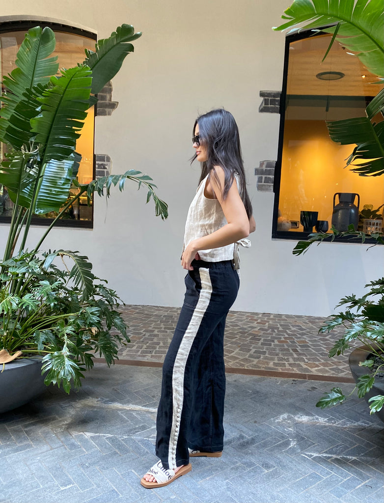 Kate Moore posing in front of some green palm leaves with our Alexa Pant in black with natural stripe down the leg and Muse Linen Vest in Natural