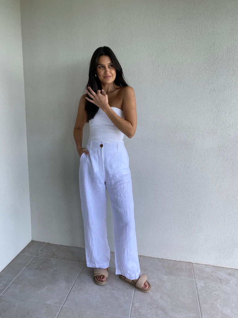 Kate Moore posing on a plain white wall wearing an al white outfit in our linen tailored pant in white and a white boob tube top and sandals