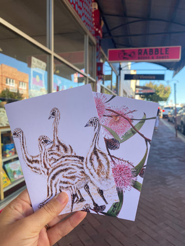 Two greeting cards held in a light skin hand with Rabble Books shop behind
