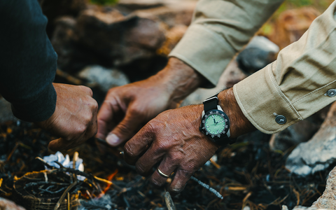 Two people start a camp fire, highlights bright green dial of watch worn