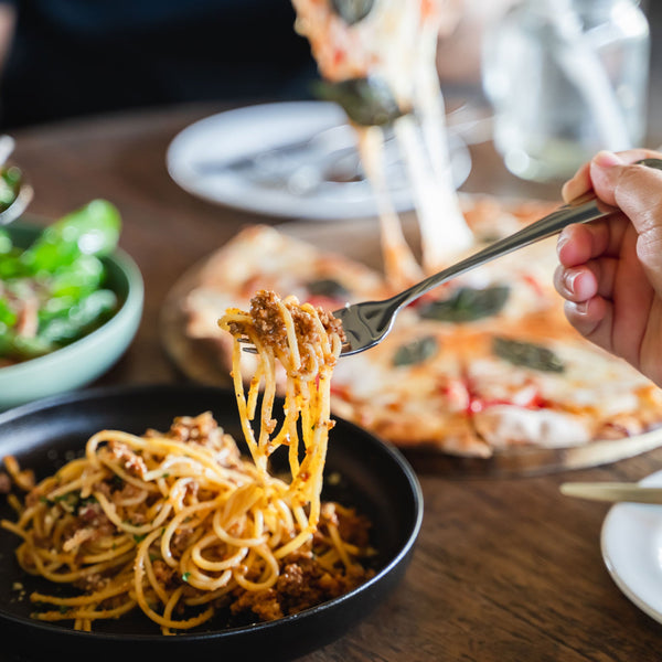 An evening dinner scene of a hand spinning spaghetti onto a fork with pizza in the background